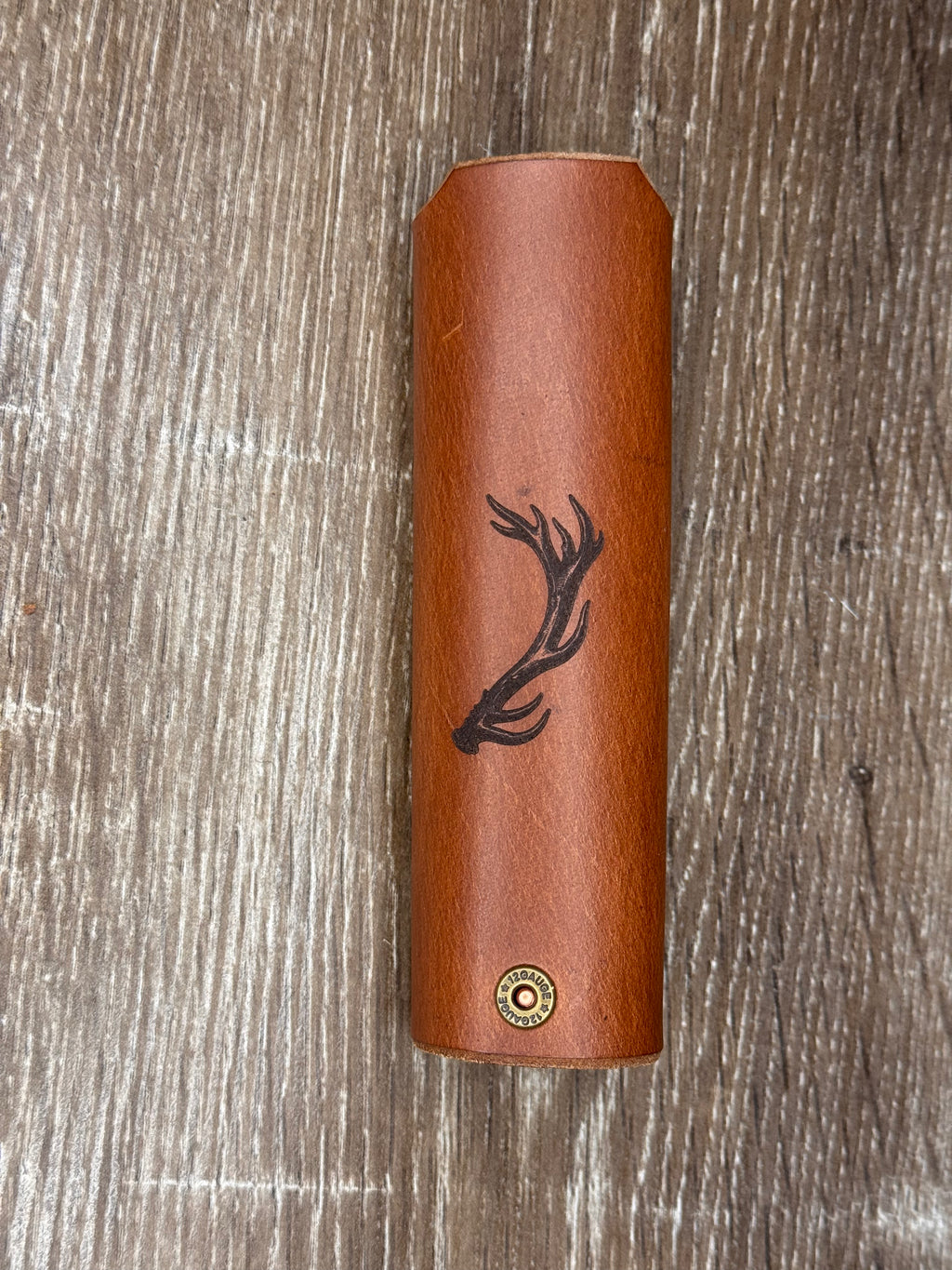 A brown leather cylindrical case with a black deer antler engraving and a small gold-toned circular emblem near the bottom, displayed on a rustic wood background.