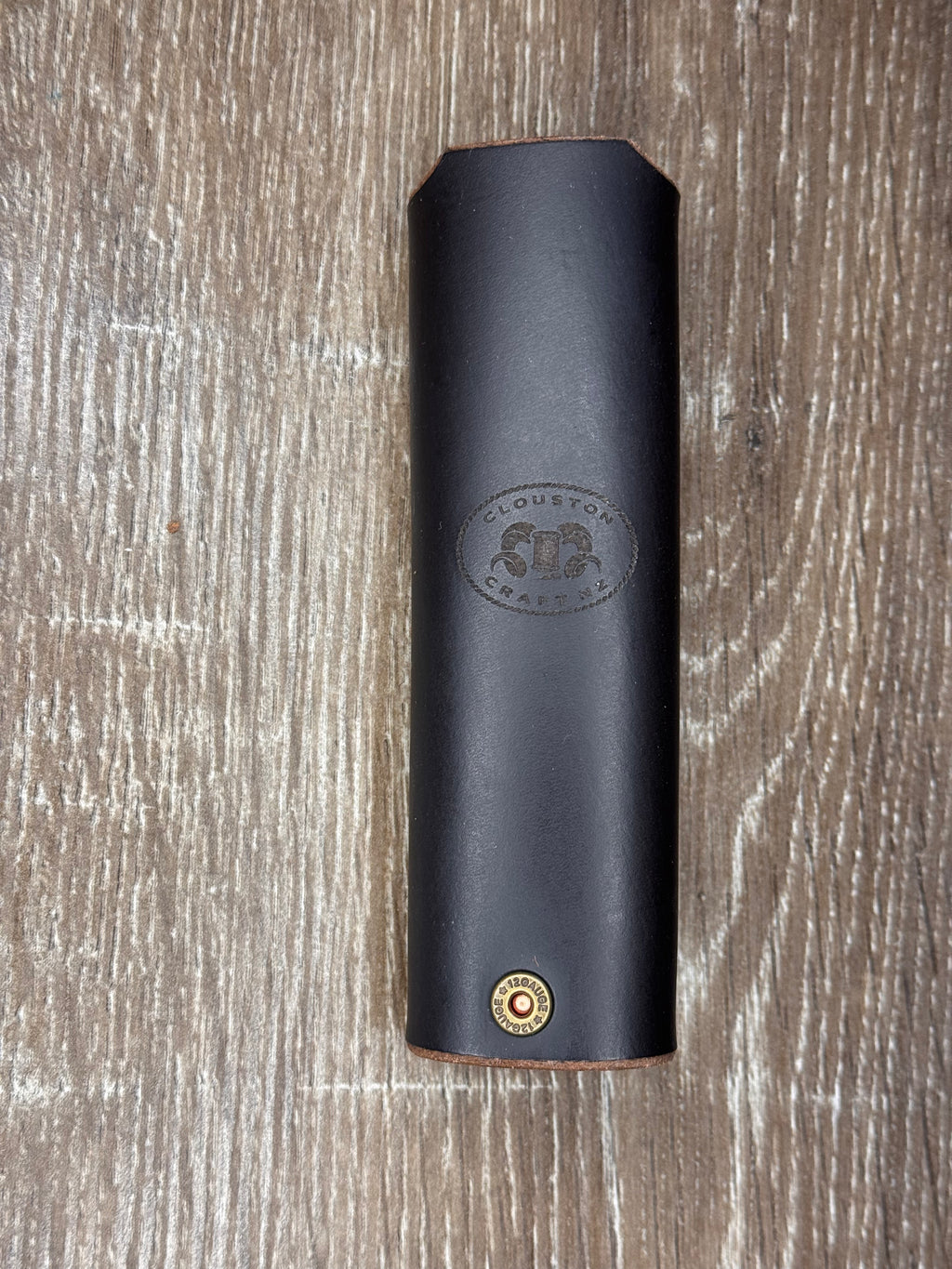 A black leather cylindrical sleeve or cover with a debossed 'Cloudstone Apparel' logo and a small brass button, shown on a brown wood background.