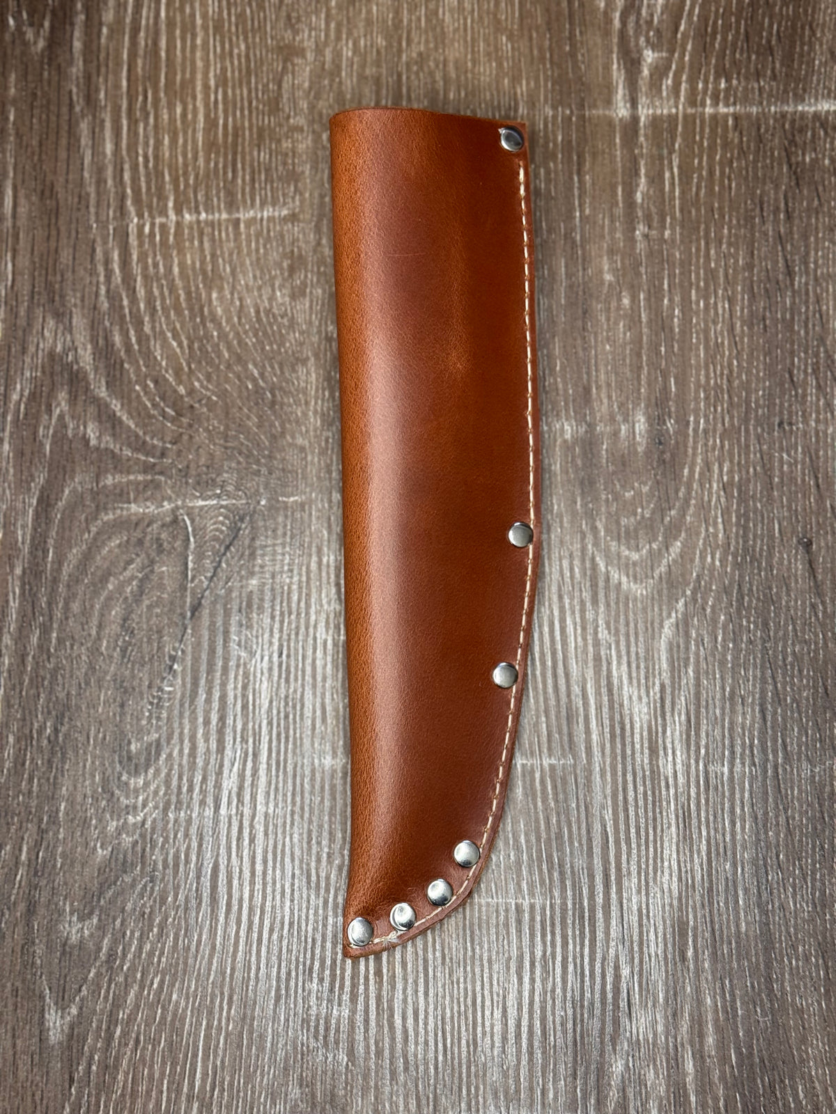 A brown leather knife sheath with silver metal rivets, laid flat on a gray-brown wood grain surface.