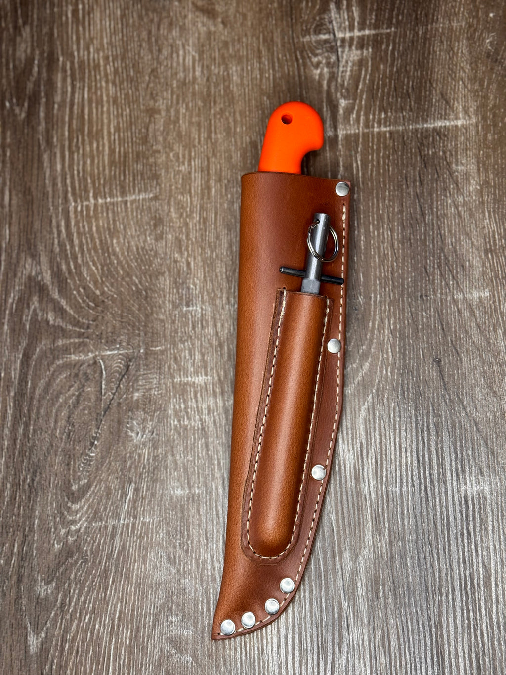 A brown leather knife sheath with silver rivets, holding a knife with a bright orange handle, placed on a gray-brown wood plank background.