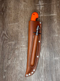 A brown leather knife sheath with silver rivets, holding a knife with a bright orange handle, placed on a gray-brown wood plank background.